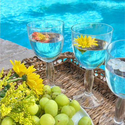 Two glasses of blue liquid with flowers, surrounded by green grapes and yellow flowers on a wicker table by a pool.